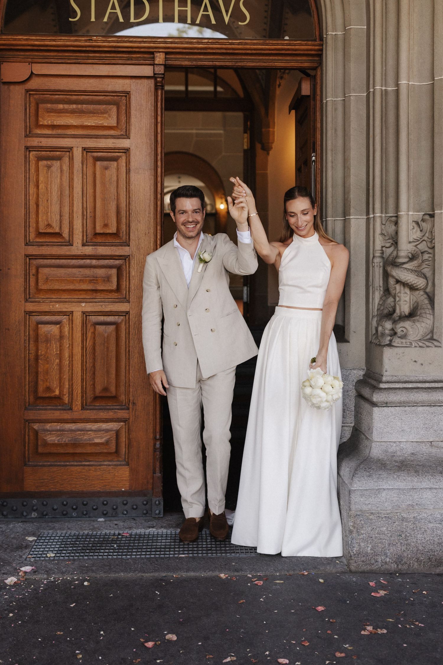 Newlyweds celebrate outside Stadthaus raising hands joyfully, bride holding white bouquet.
