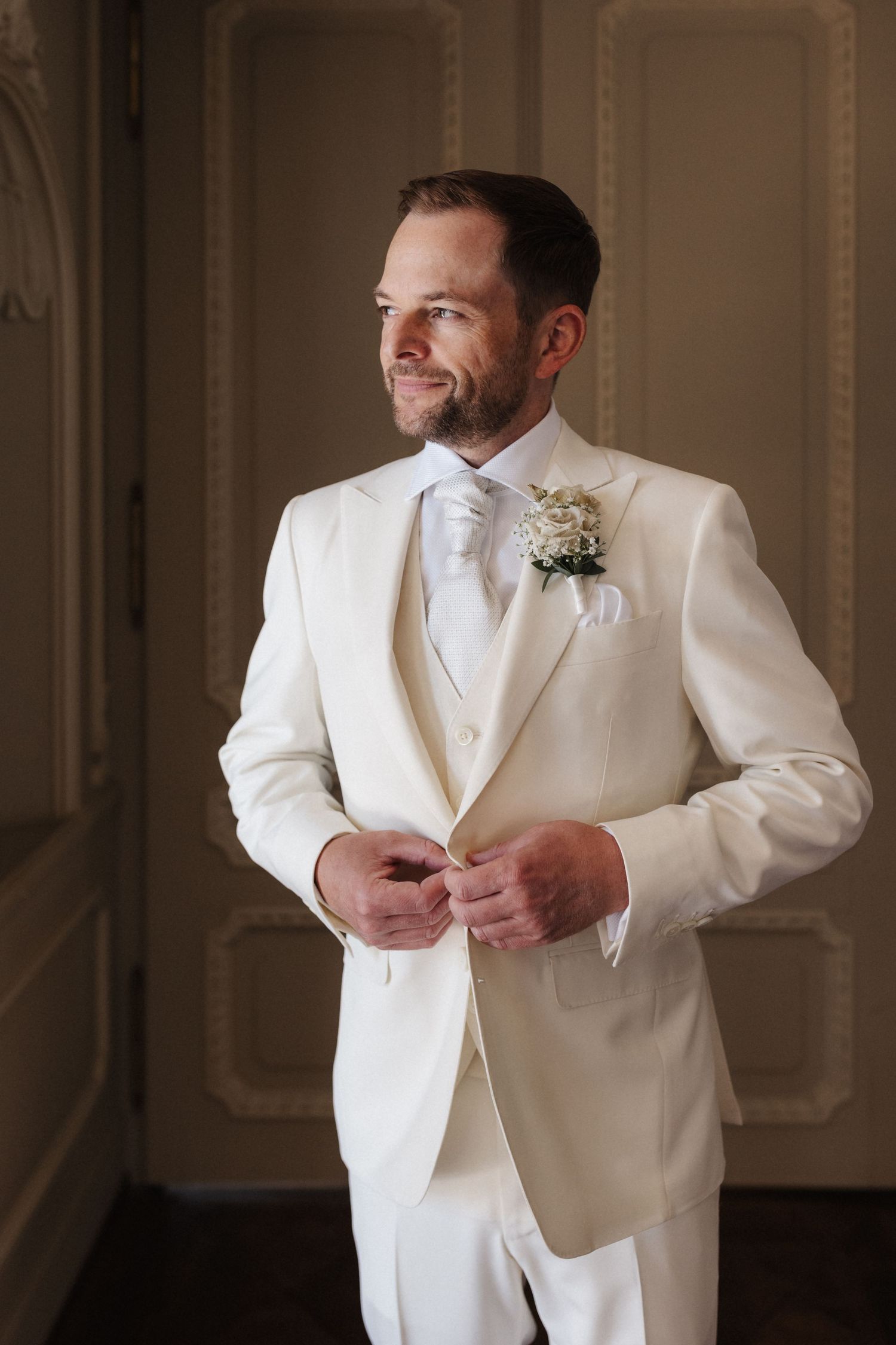 Dapper groom in ivory three-piece suit with floral boutonniere buttoning jacket before wedding.