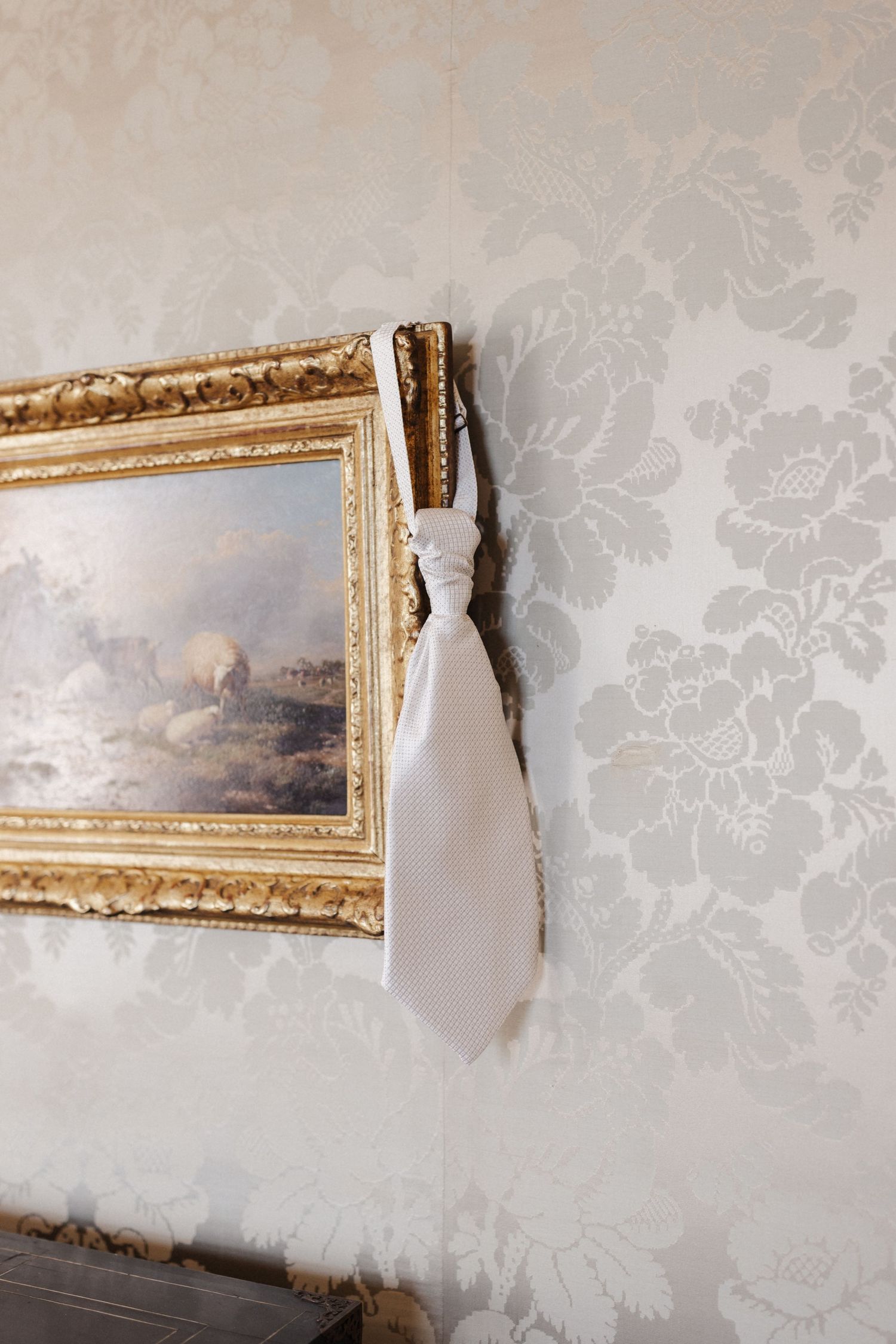 White cravat tie hanging on ornate gold picture frame against elegant floral damask wallpaper.