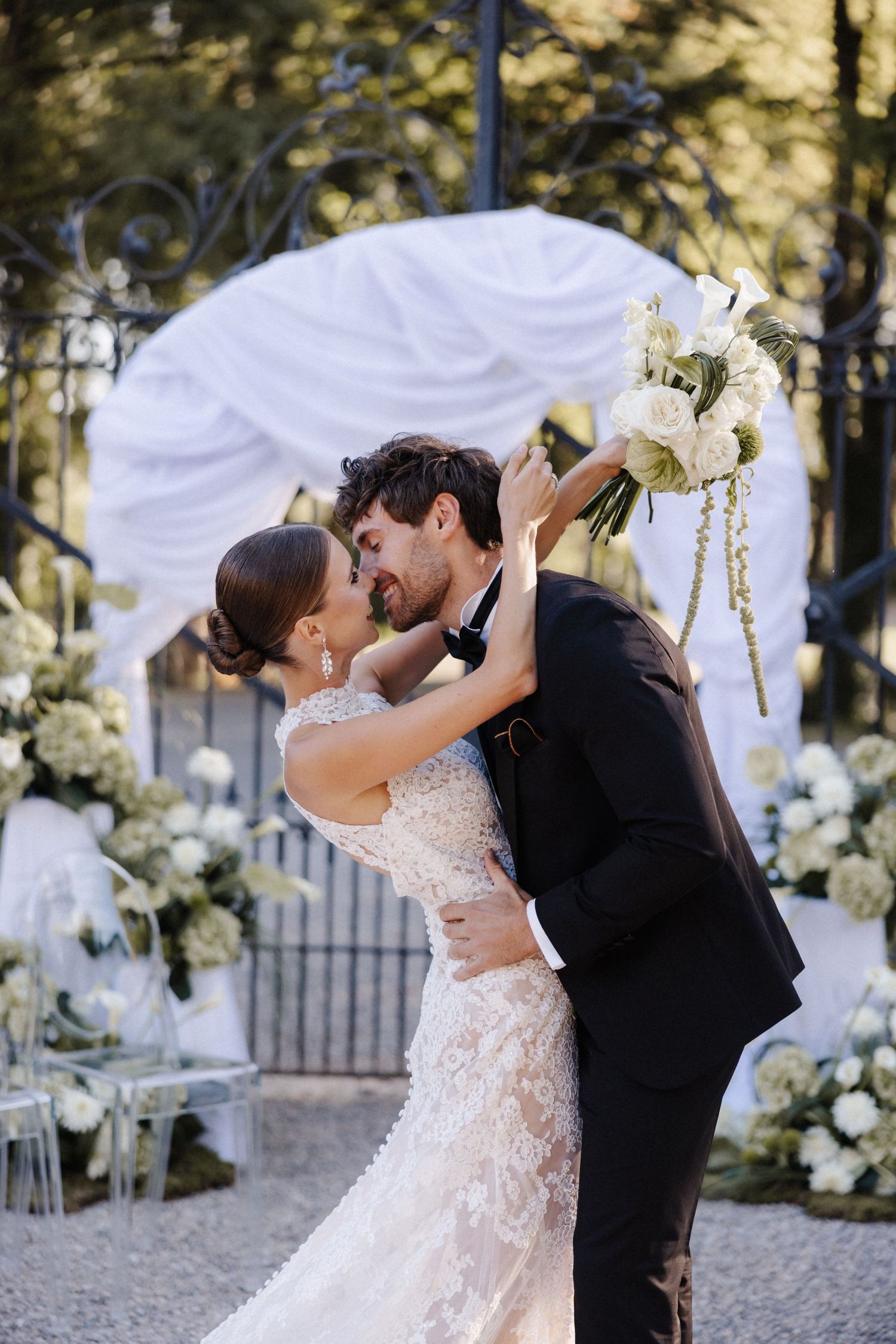 Groom dips bride near a white floral arch as she holds a bouquet of white flowers outdoors.