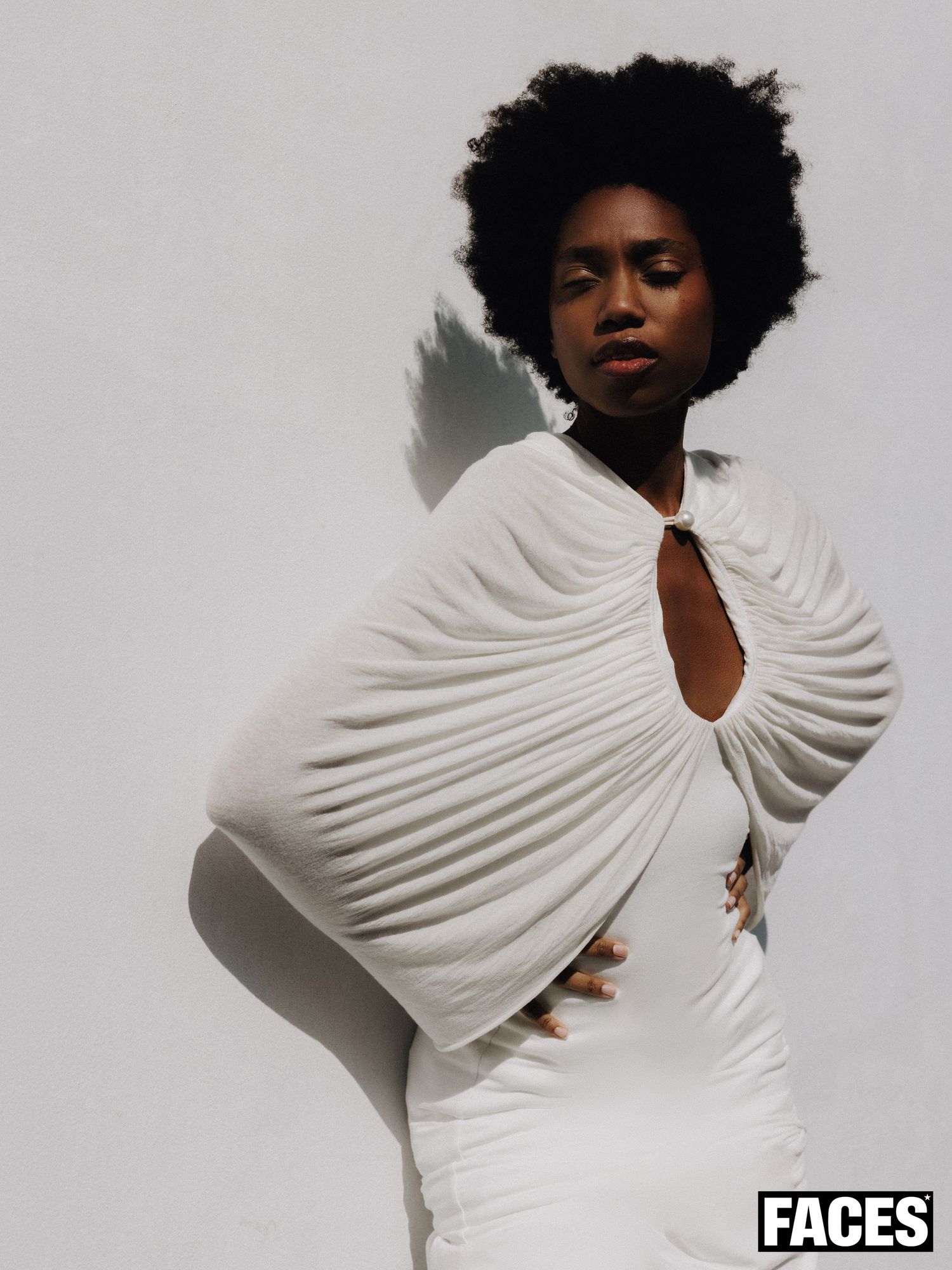 A Black woman with an afro poses in a white pleated cape dress with a keyhole neckline against a white wall.