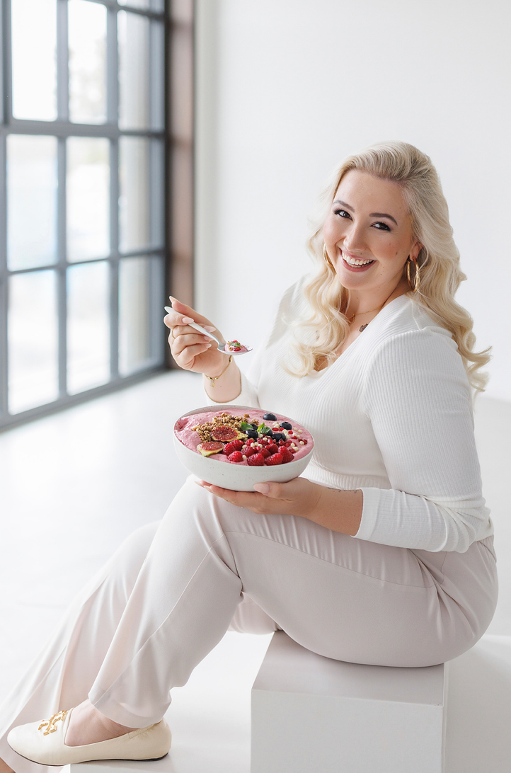 Smiling blonde woman in white outfit holding a colorful berry smoothie bowl with a spoon.