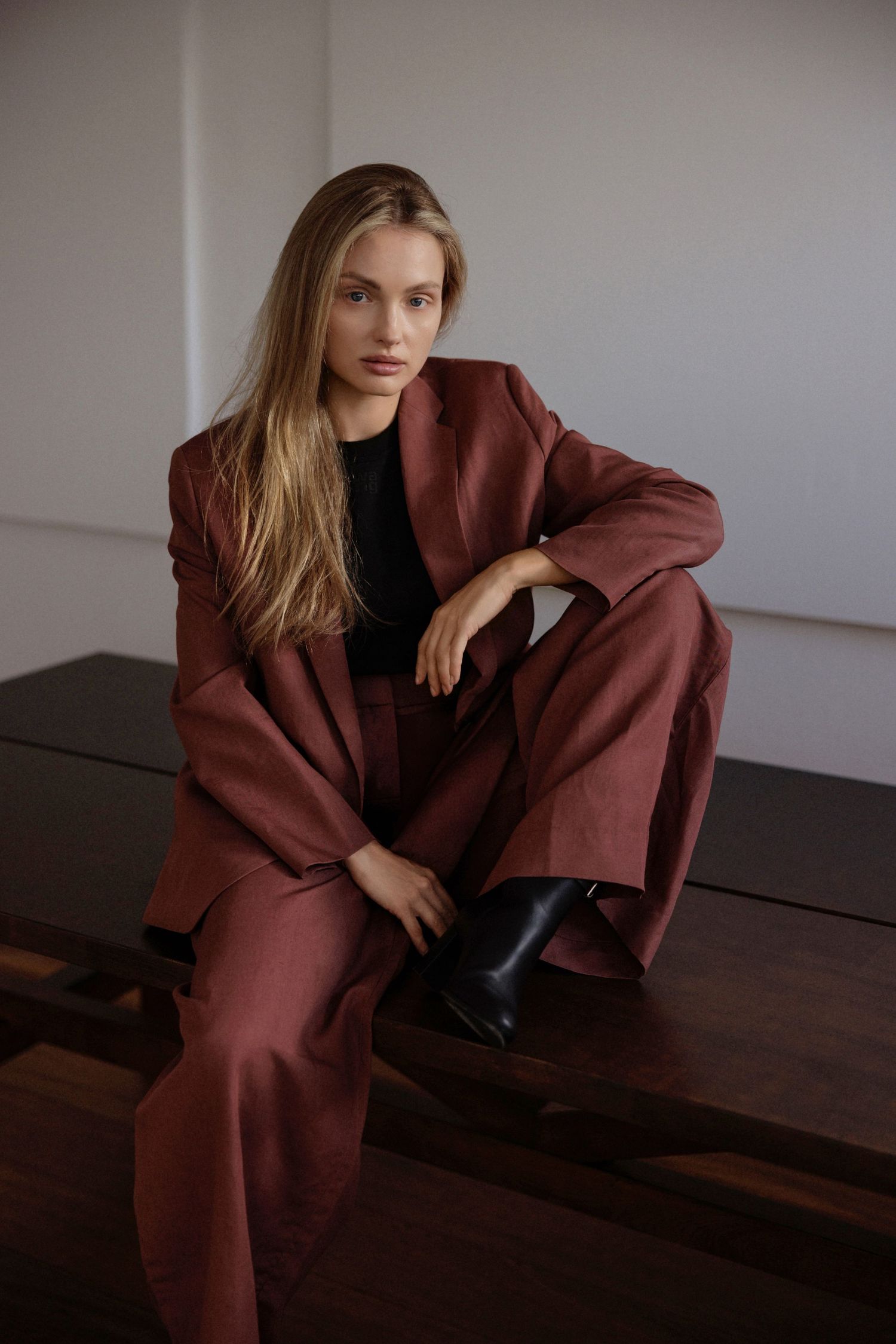 Woman with long blonde hair sits on a dark wooden bench wearing a rust-brown linen blazer, wide-leg trousers, black top and black ankle boot