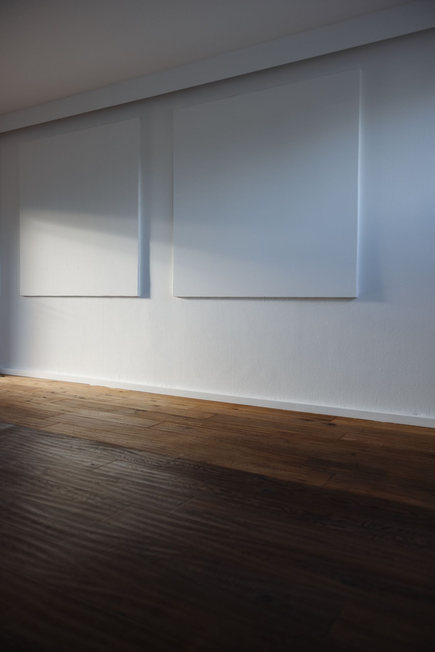Two white canvas artworks hanging on a gray wall above a dark hardwood floor in a minimalist gallery.
