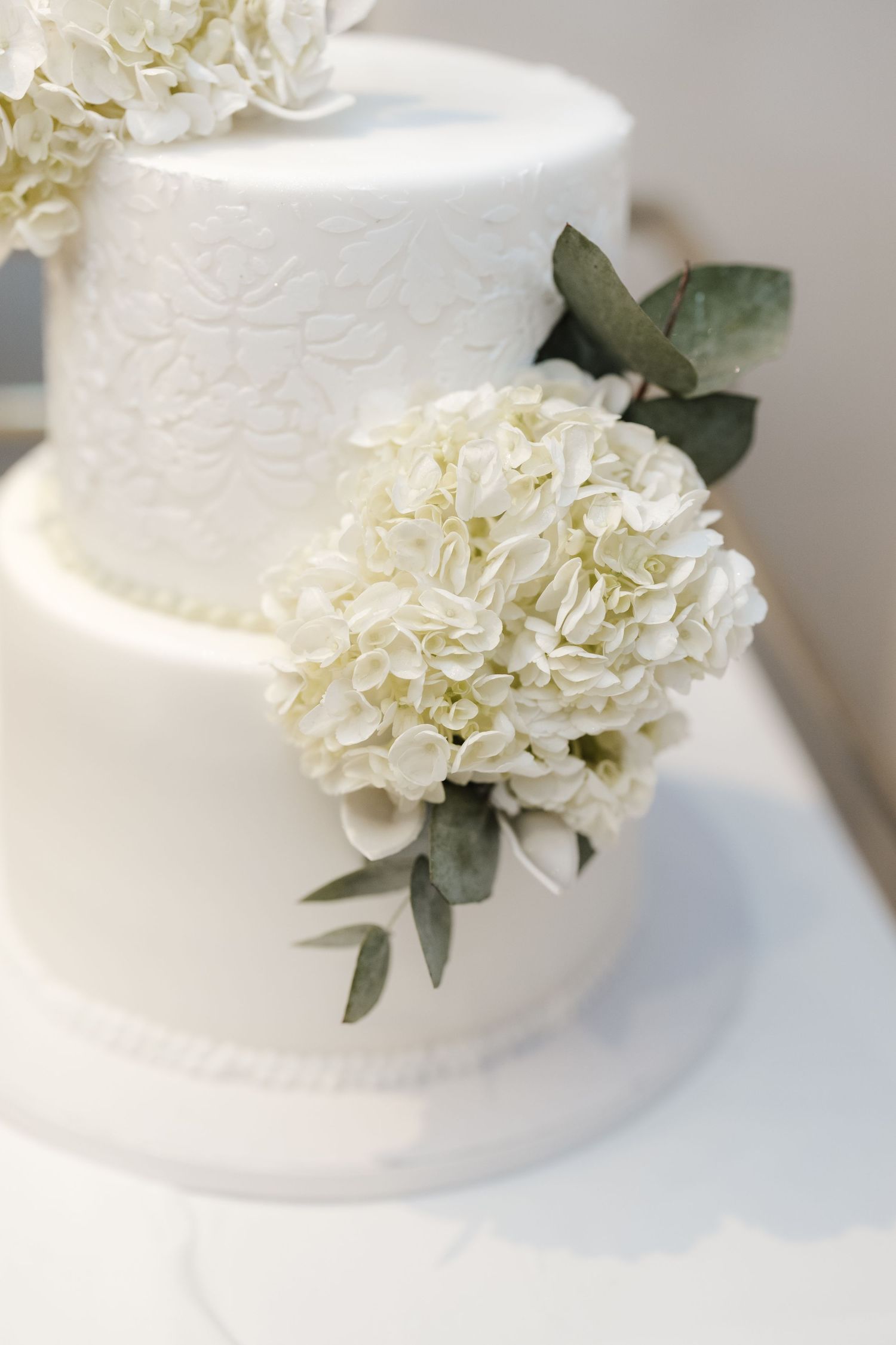 Elegant white wedding cake with hydrangea flowers and eucalyptus leaves on a white fondant tiered design.