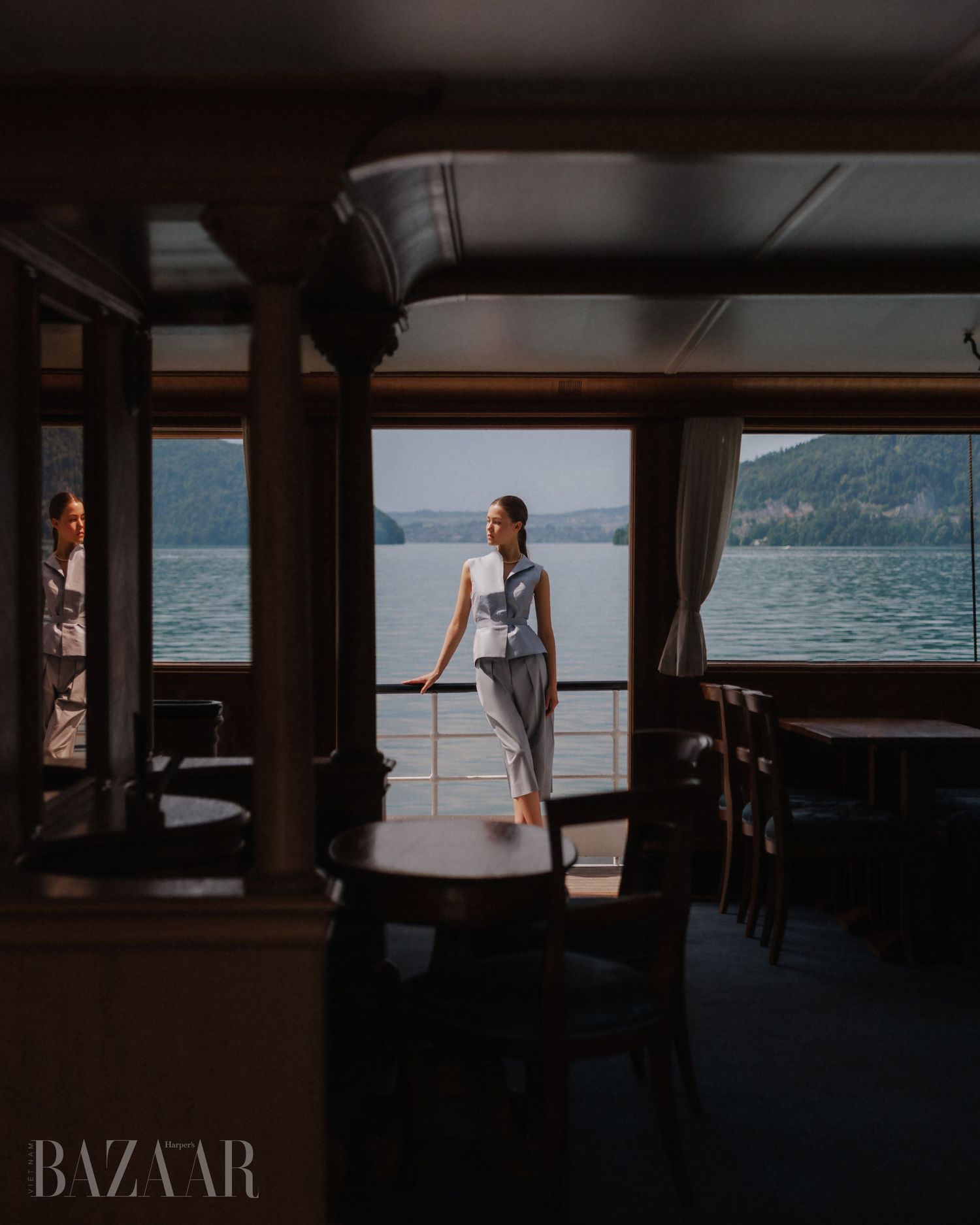 Model in blue ensemble standing on boat deck with panoramic lake and mountain view for Harper's Bazaar editorial.