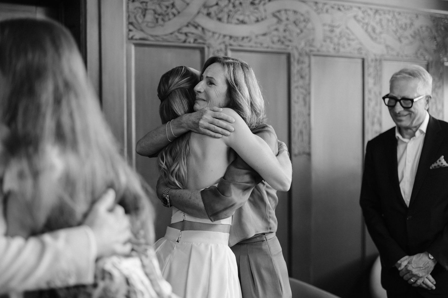 Black and white photo of bride hugging female guest while older man smiles warmly in background at wedding reception.