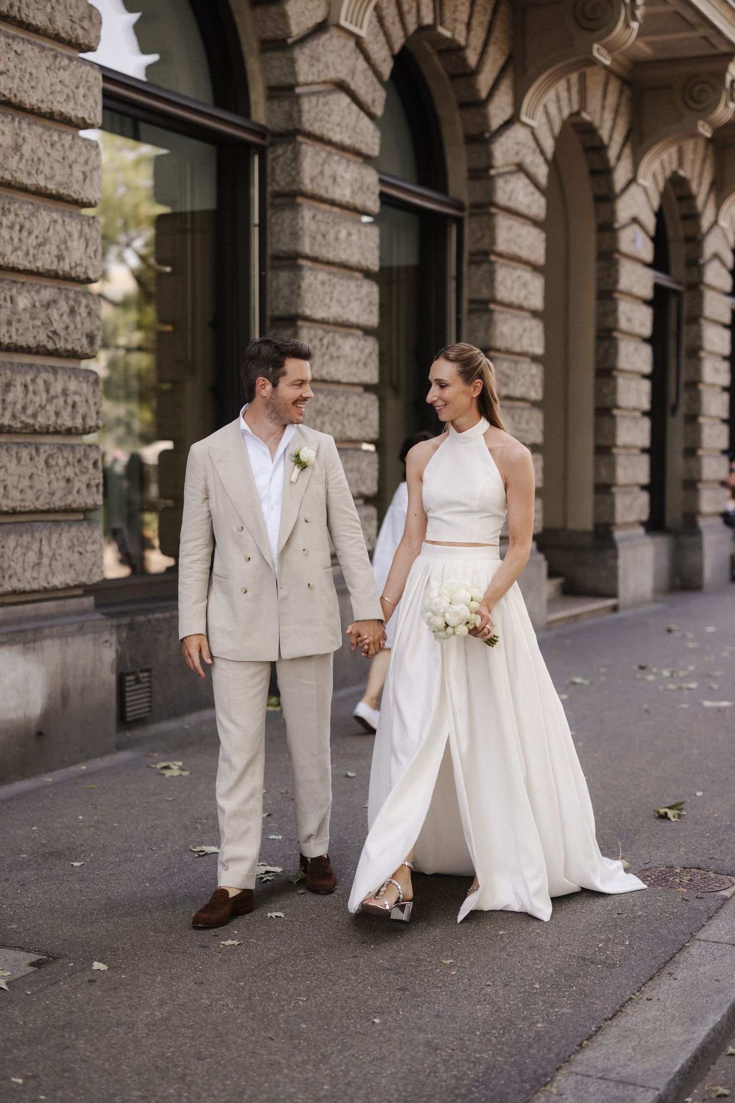 Elegant bride and groom walk hand in hand along city street in matching neutral wedding attire.