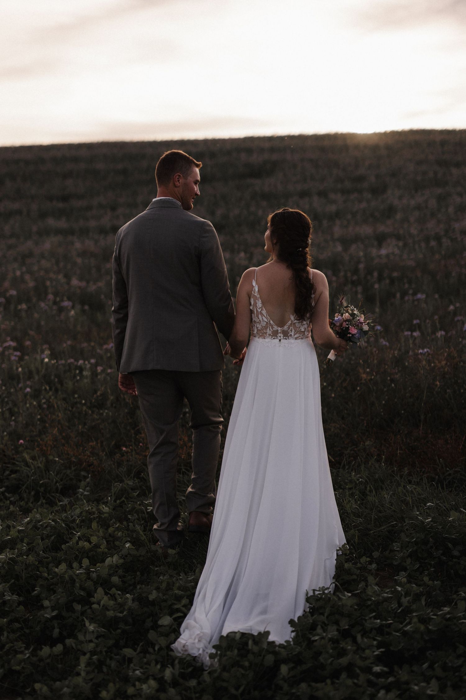 Bride and groom hold hands facing a stunning sunset over a wildflower field on their wedding day.