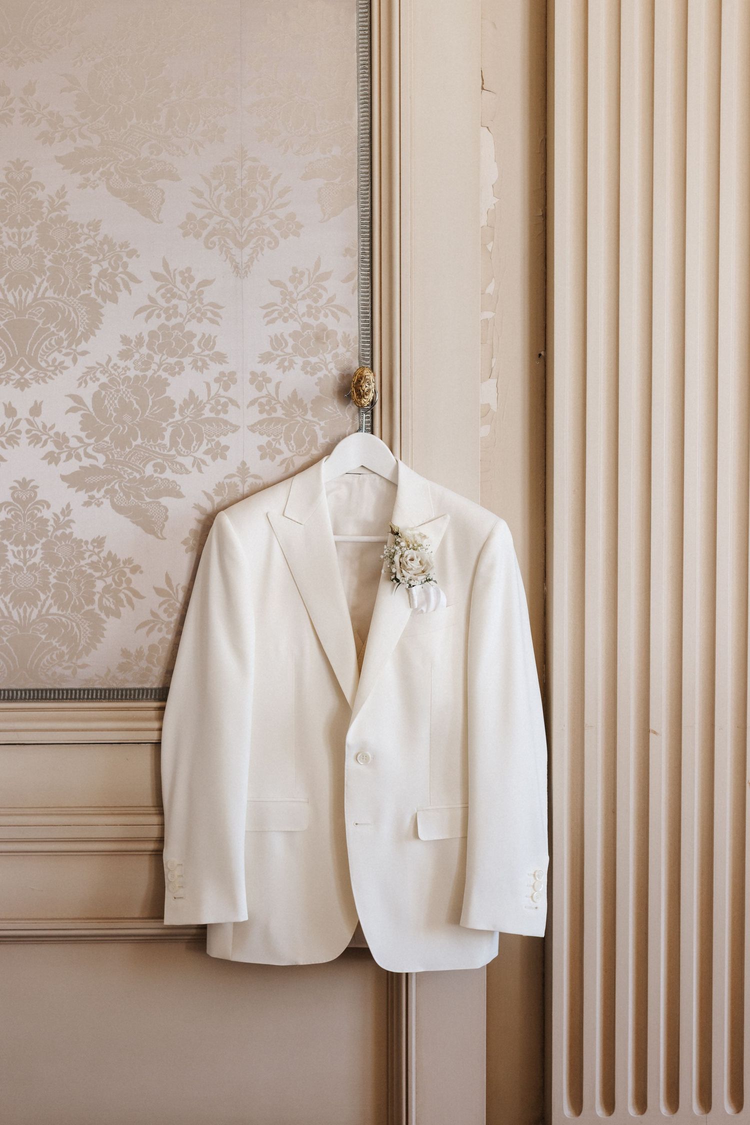 White wedding suit jacket with floral boutonniere hanging on ornate gold hook against damask wallpaper.