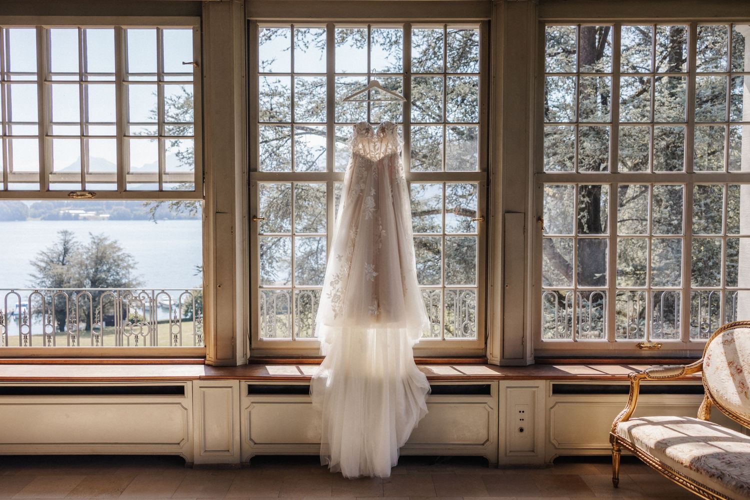 An elegant lace wedding dress with a long veil hanging in front of large vintage windows.