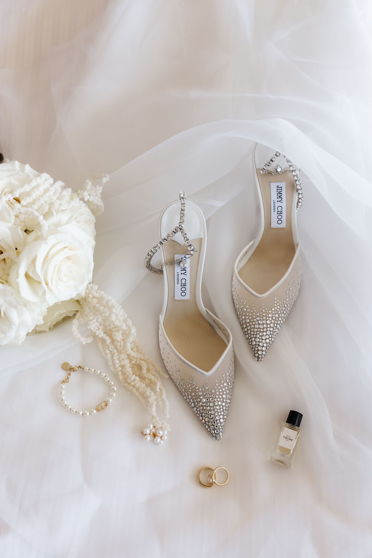 Jimmy Choo crystal embellished bridal heels with pearl necklace, rings, and perfume on white tulle.