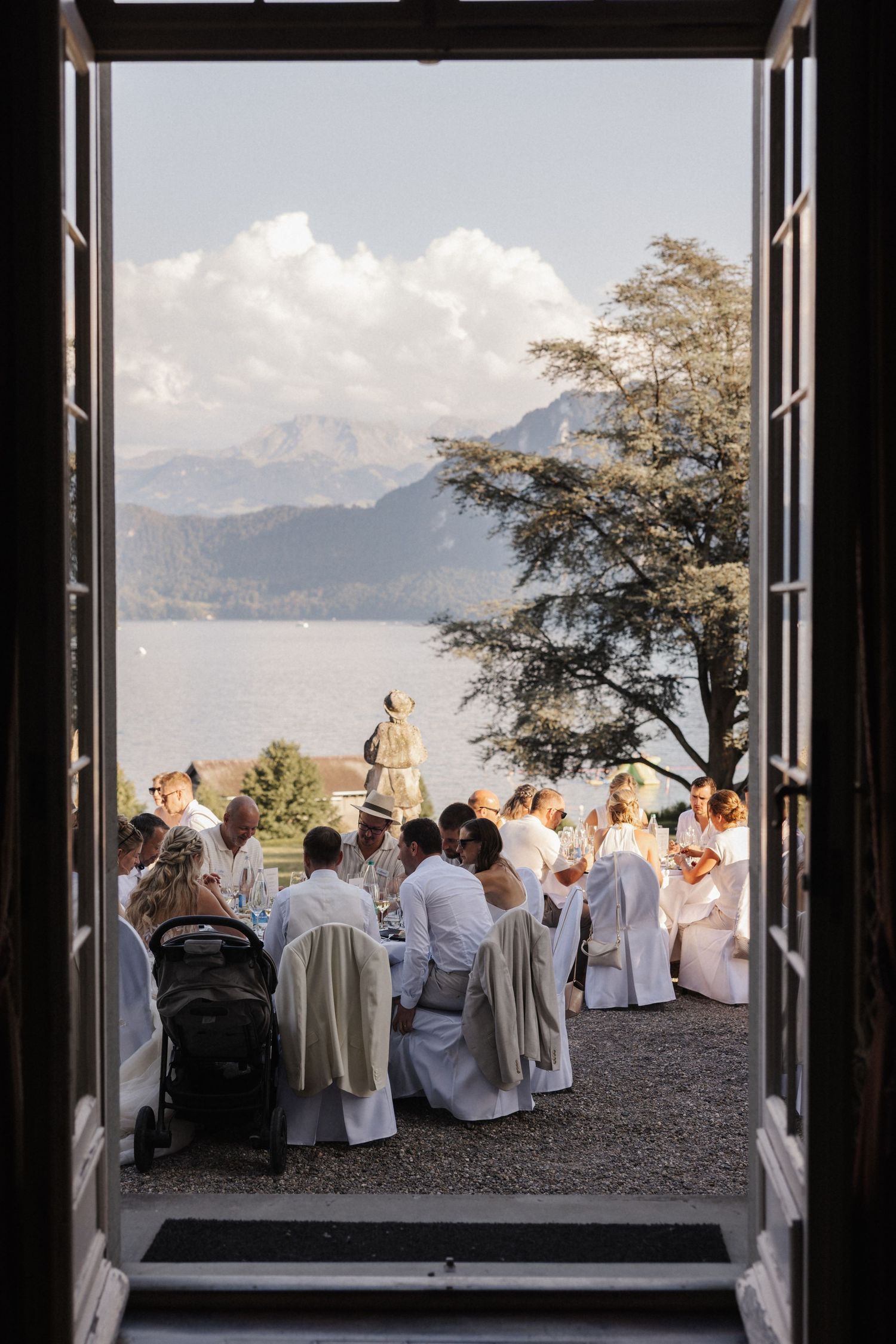 Wedding reception viewed through open French doors with guests in white seated overlooking a serene mountain lake.