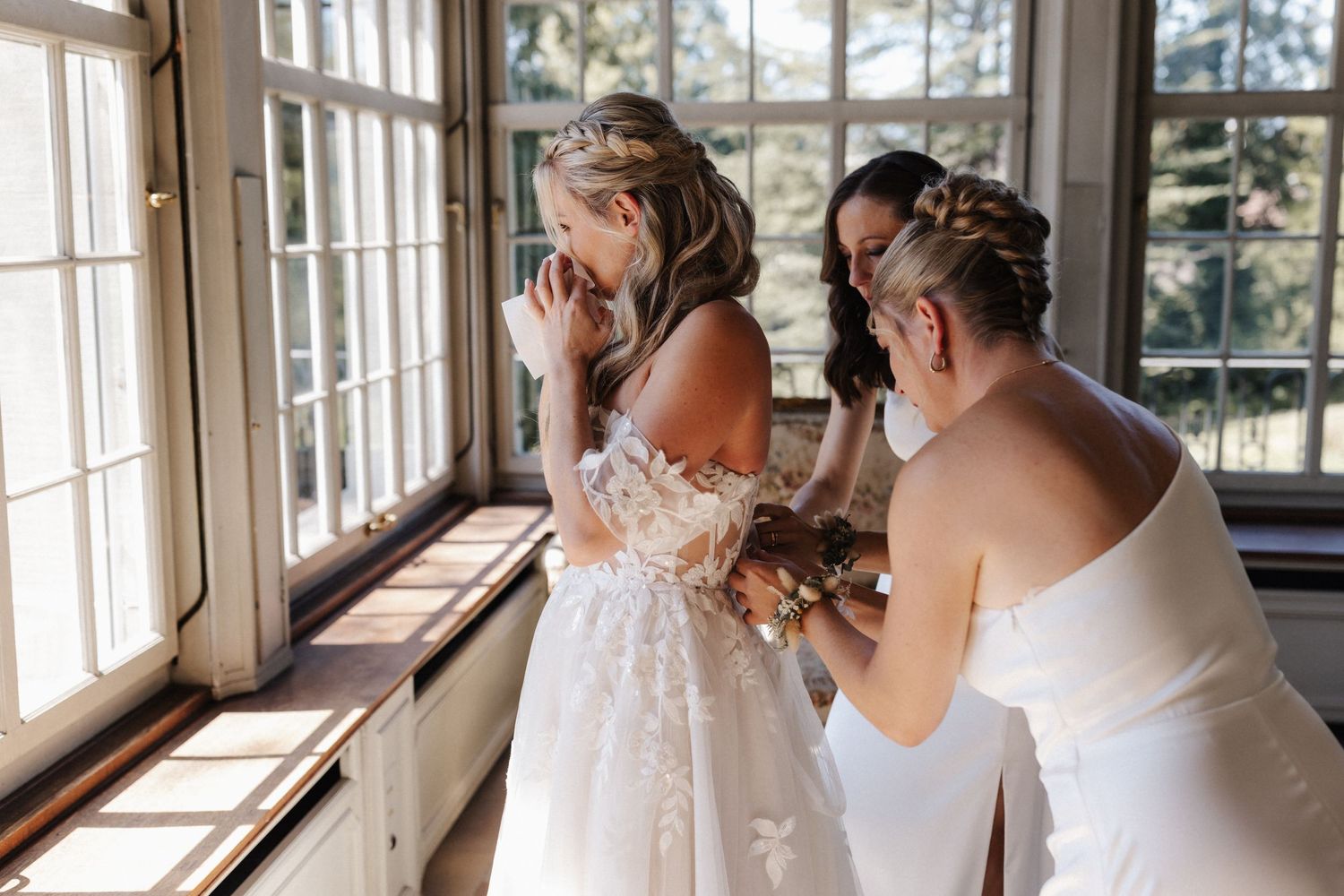 Emotional bride in a lace gown being comforted by two bridesmaids near sunlit windows.