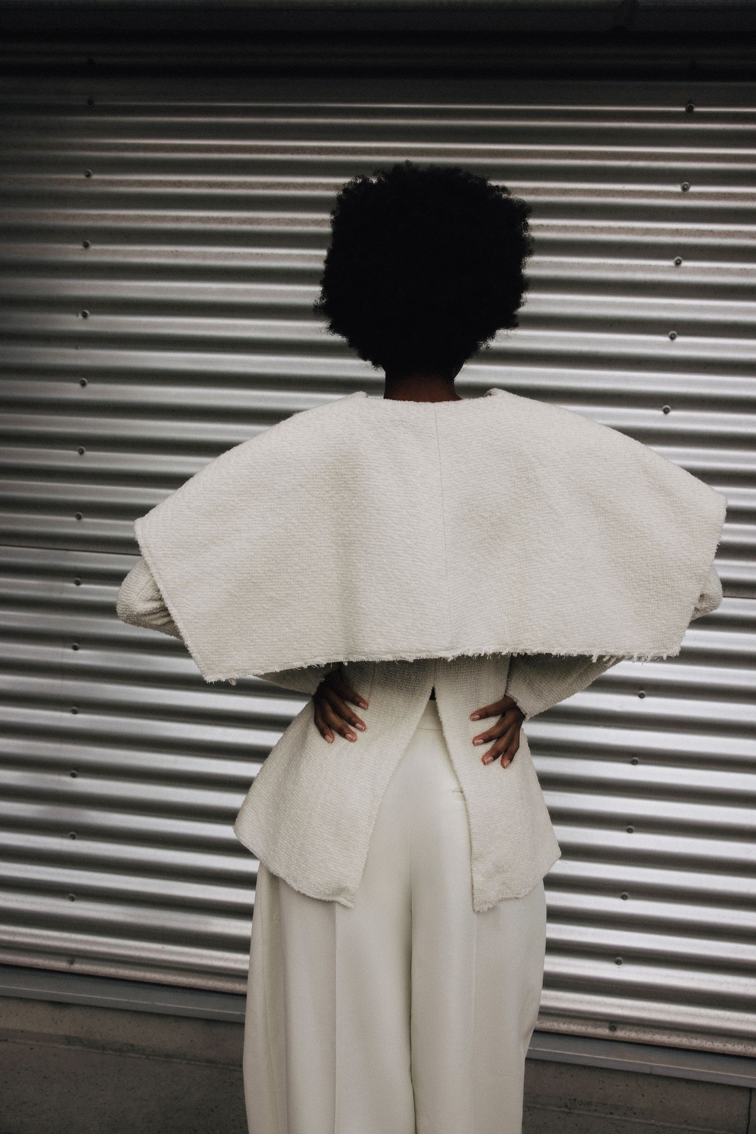 Back view of model wearing a wide white boucle cape over white trousers against a corrugated metal backdrop.