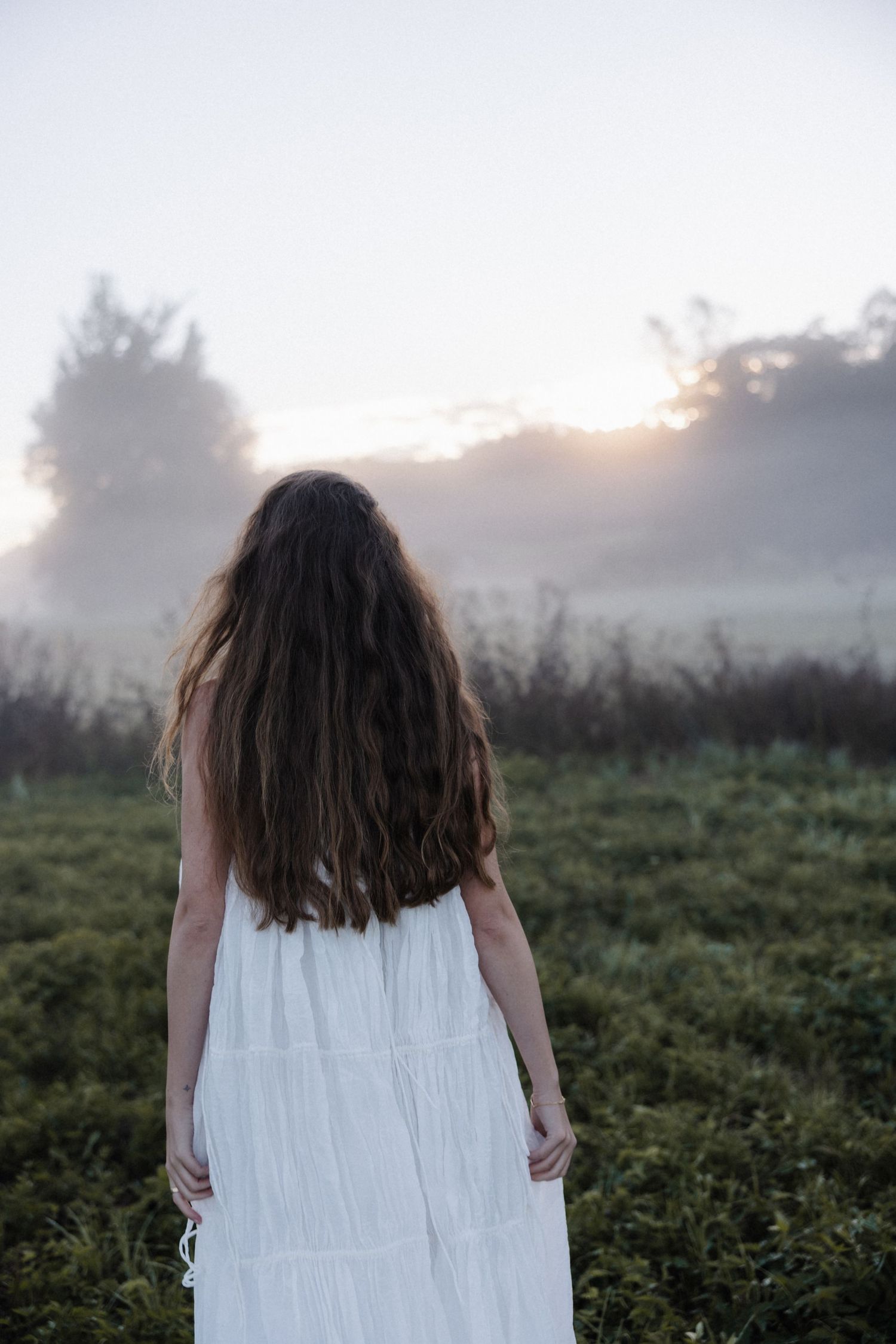 Woman with long curly hair in white dress standing in misty field at sunrise viewed from behind.