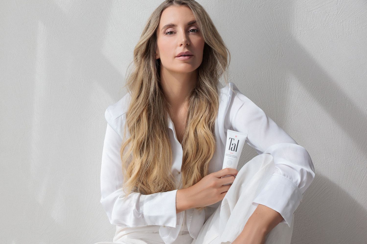 Blonde woman in white outfit sitting against a gray wall holding a Tal skincare product tube.