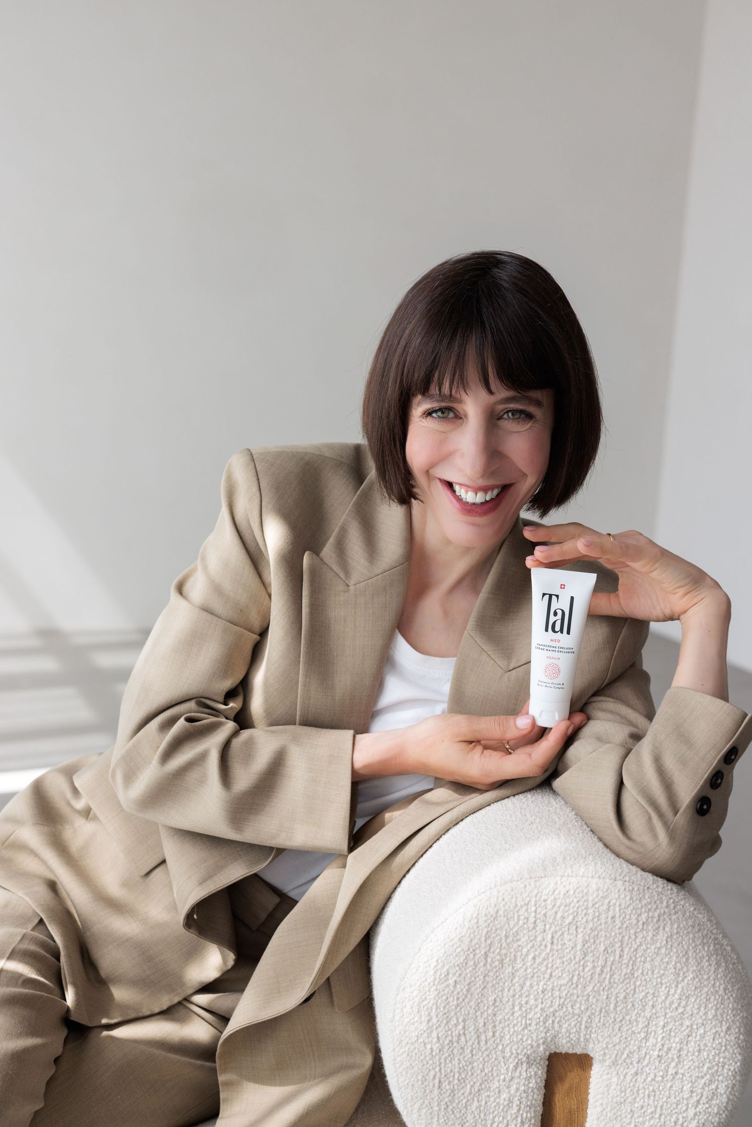 Smiling brunette woman in beige blazer holding Tal skincare product tube while seated indoors.