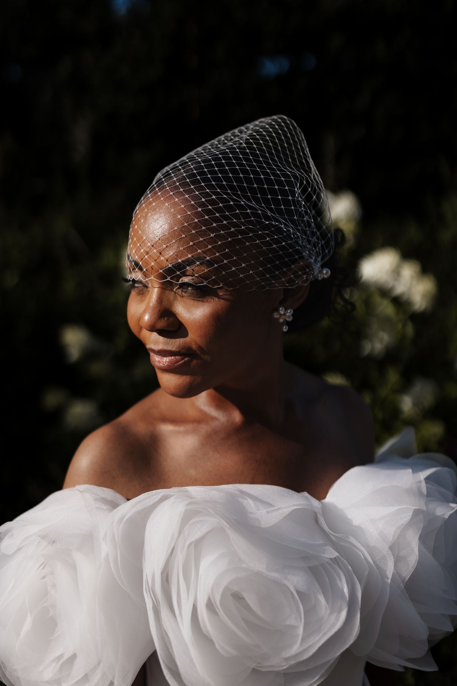 Portrait of a radiant bride wearing a birdcage veil, floral earrings, and an off-shoulder ruffled white gown.