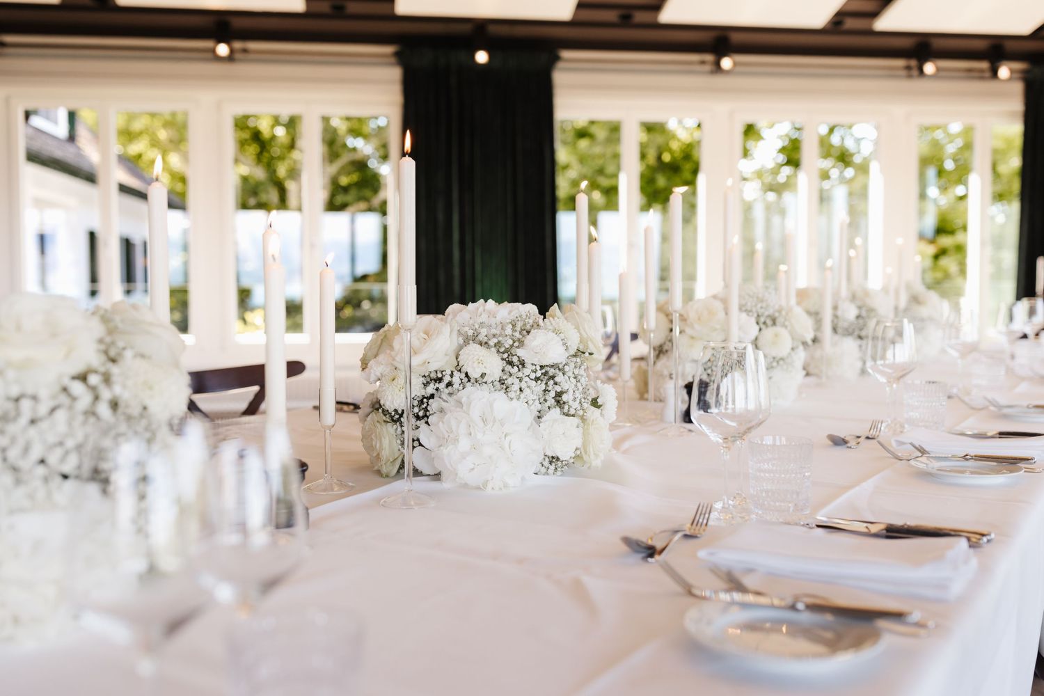 Sophisticated wedding table setting with white floral centerpieces, tall taper candles, and elegant place settings in bright venue.
