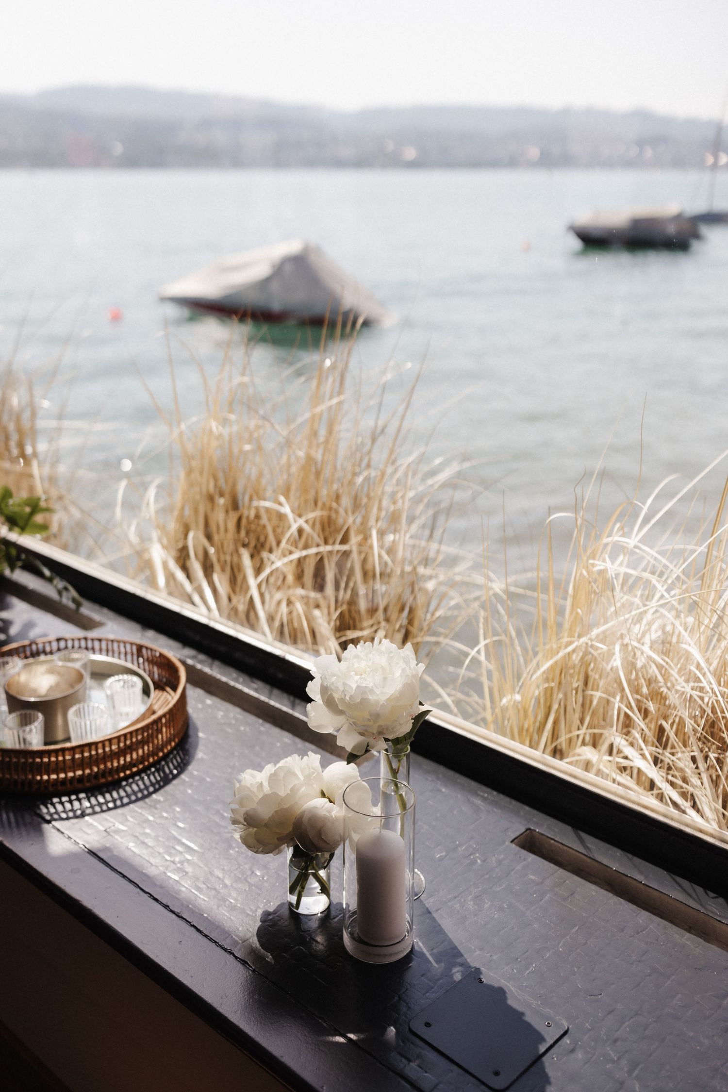 White peony flowers in a vase on a dark surface overlooking a serene lake with boats.