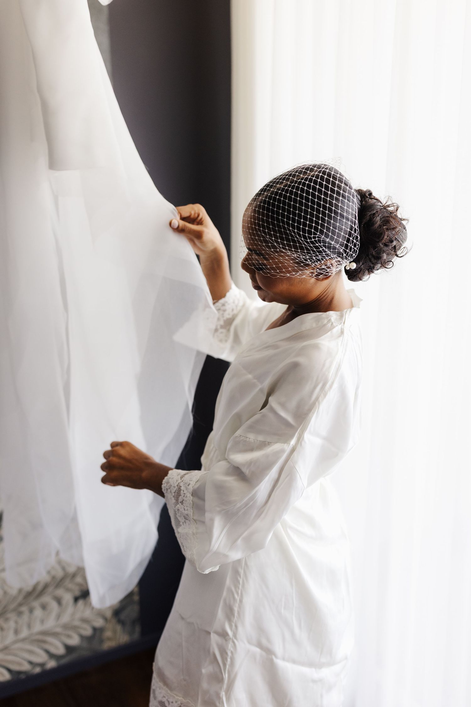 Black bride in white robe and birdcage veil reaching for her wedding dress hanging by window.