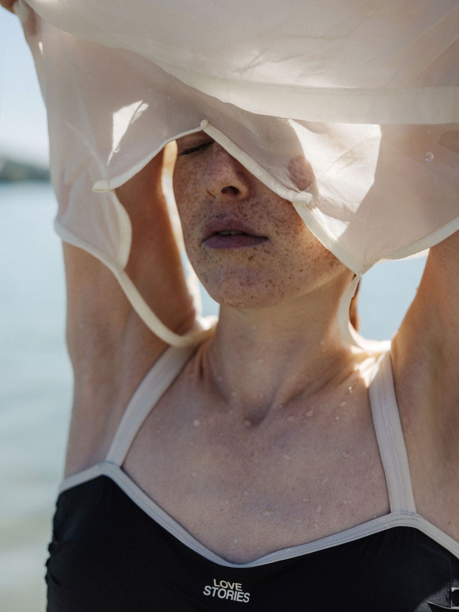 Woman in black Love Stories swimsuit with white fabric draped over her face near water.