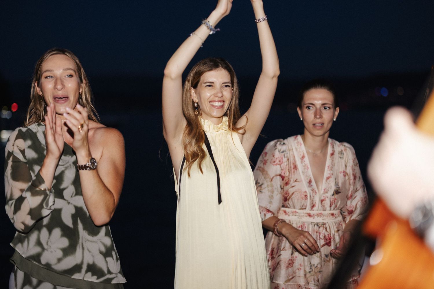 Three women celebrating at an outdoor evening wedding reception with arms raised.