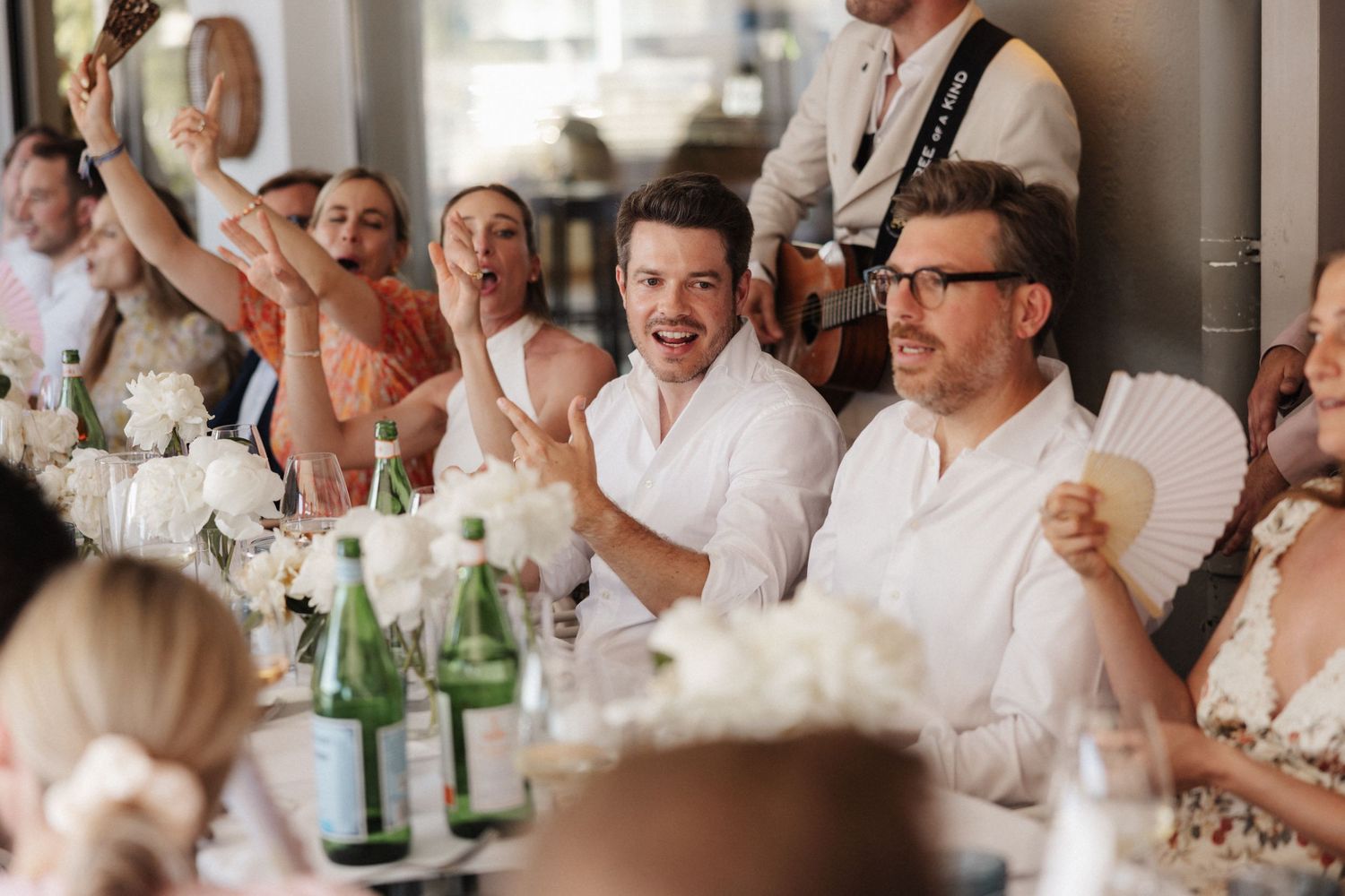 Excited wedding guests laugh and wave fans at a reception table with green glass bottles.