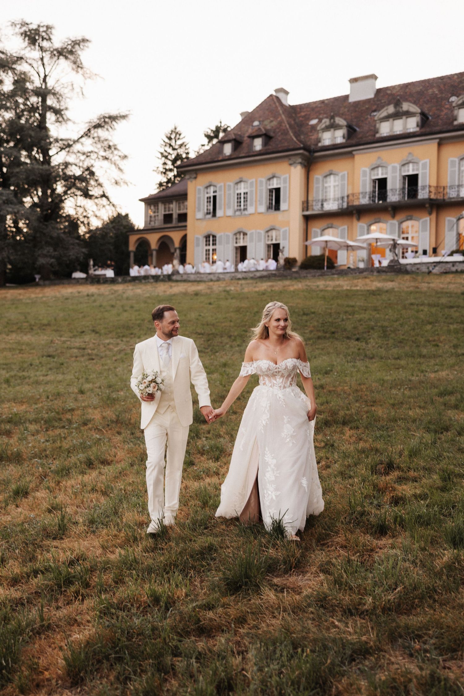 Bride in off-shoulder lace gown and groom in cream suit walking hand-in-hand across a lawn at sunset.