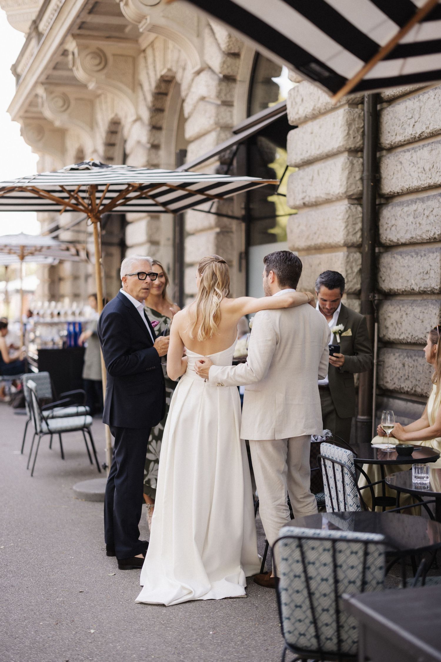 Bride and groom embrace with guests at chic outdoor cafe wedding reception venue.