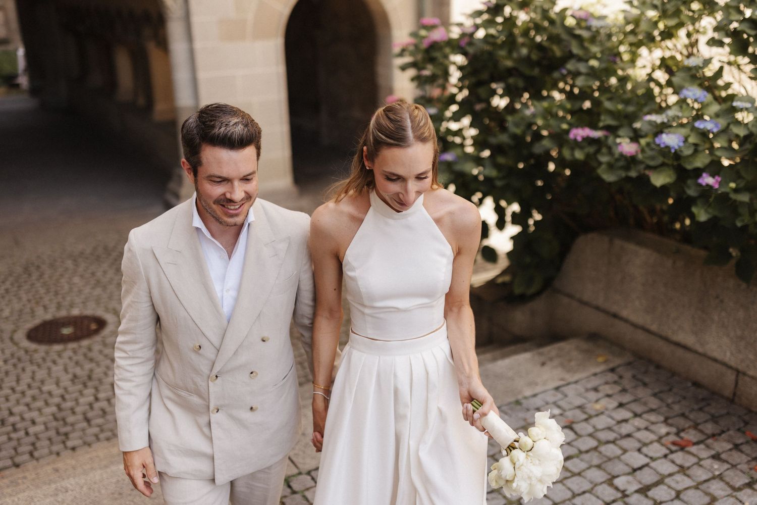 Bride and groom walking together on cobblestone street, she holds white peony bouquet, both smiling in elegant wedding attire.
