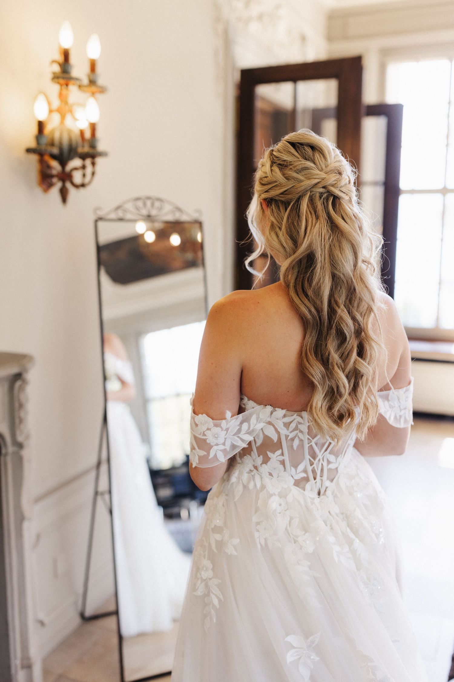Back view of bride in off-shoulder lace gown with flowing curled hair standing before a mirror.