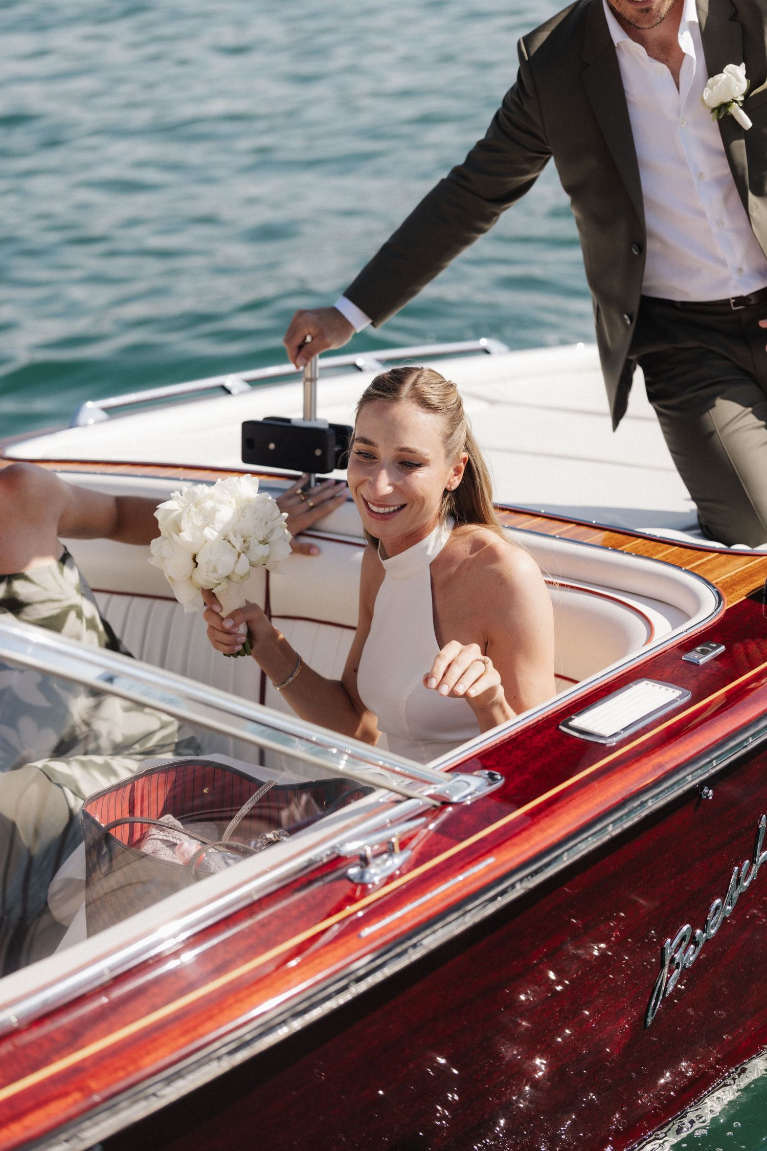 Smiling bride in halter wedding dress sitting in a classic wooden speedboat holding white bouquet.