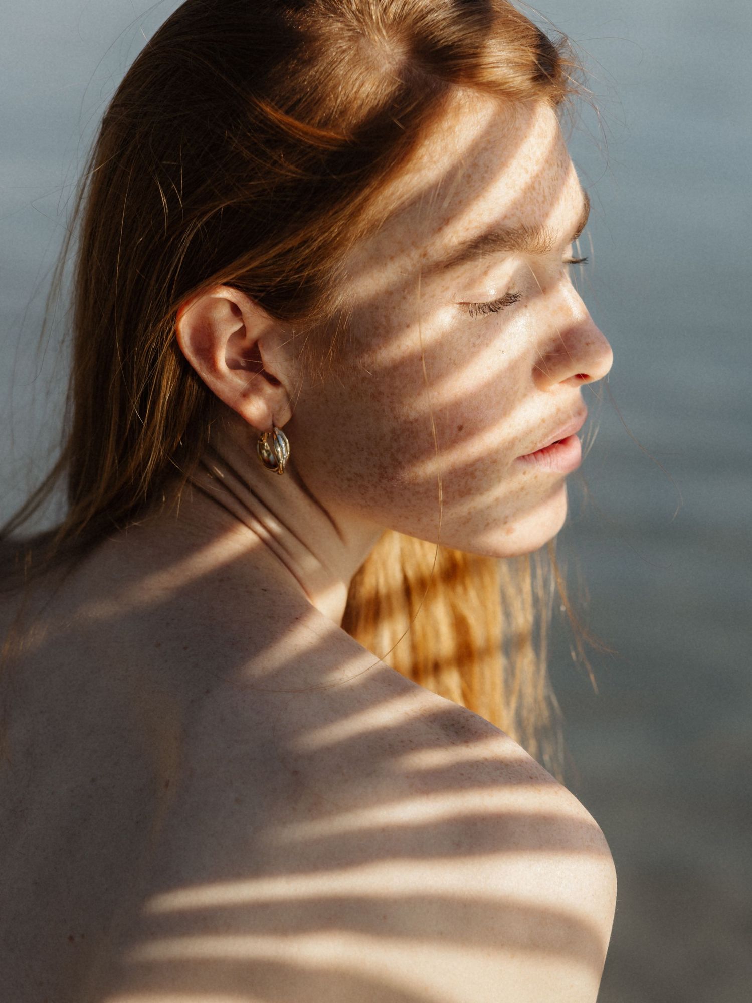 Profile of freckled woman with striped shadows across her face and bare shoulder.
