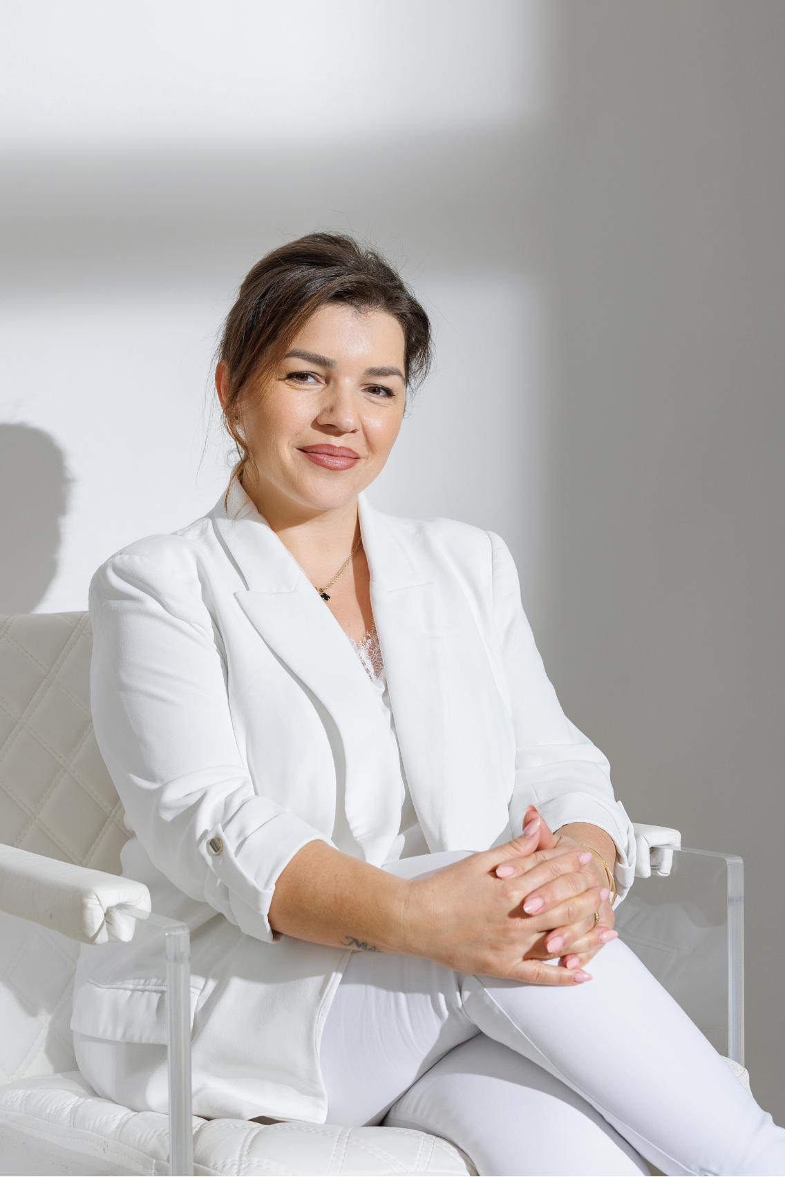 A smiling woman in a white blazer sits confidently in a clear chair against a light gray background.