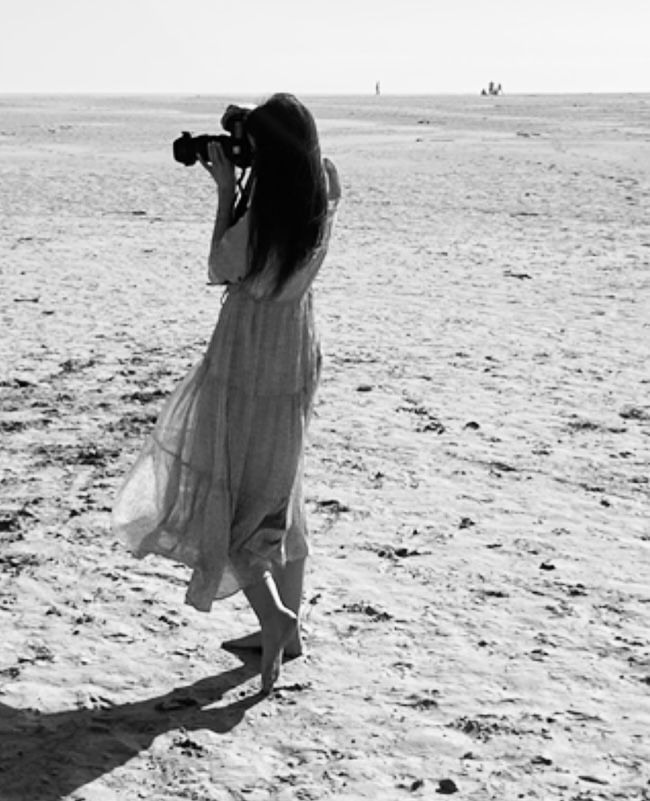 Black and white photo of barefoot woman in flowy dress photographing vast sandy beach.