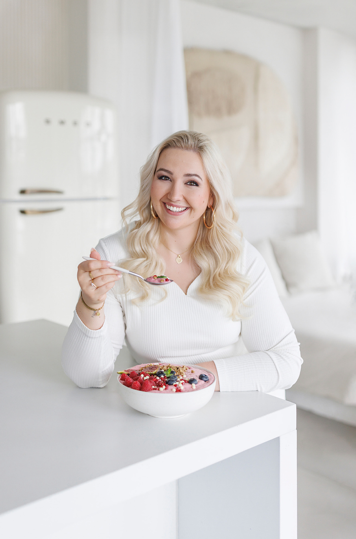 Smiling blonde woman in white top holding a berry smoothie bowl at a white kitchen counter.