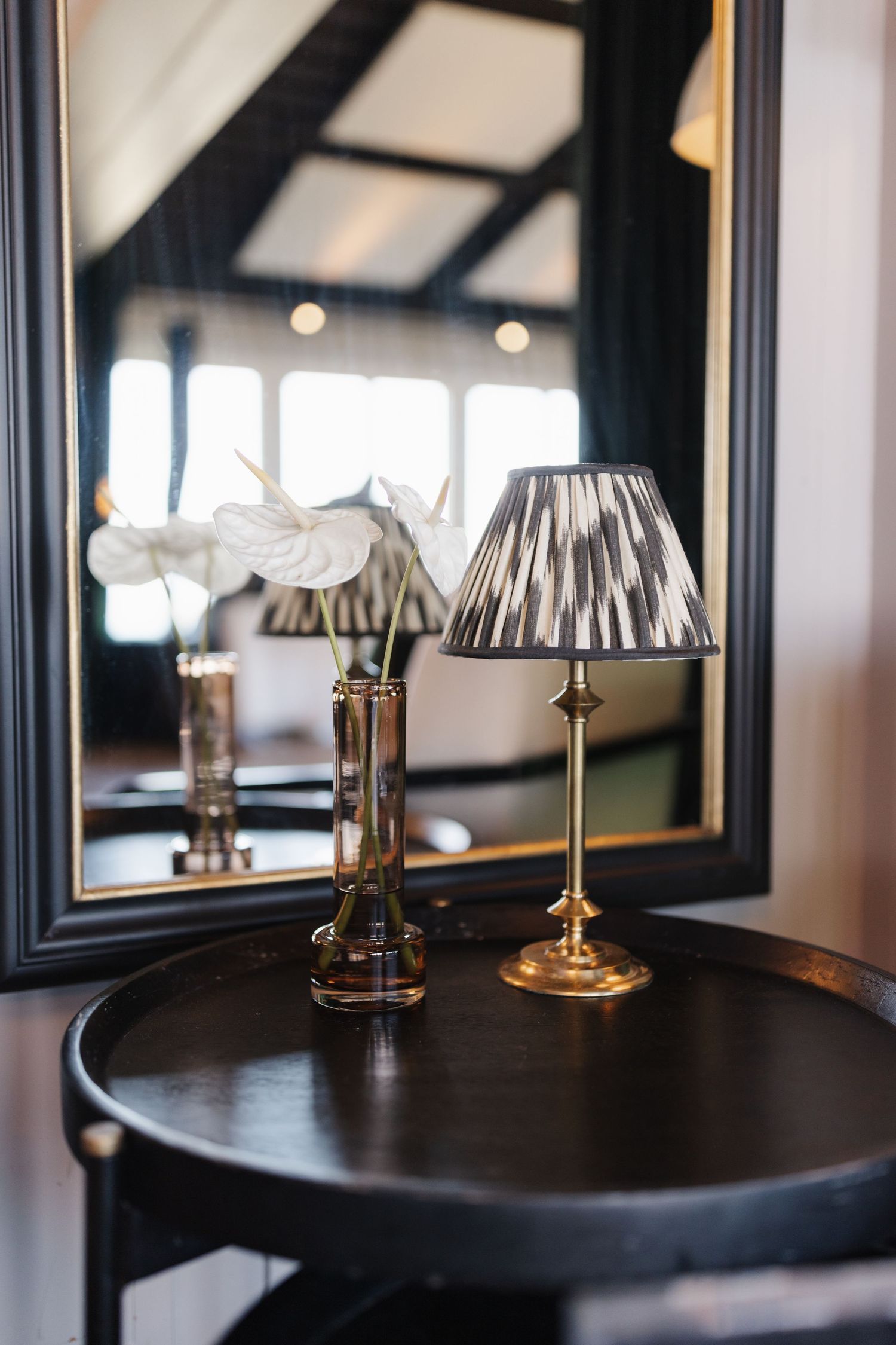 Dark wooden side table with gold brass lamp, white anthurium flowers in vase, and decorative mirror.