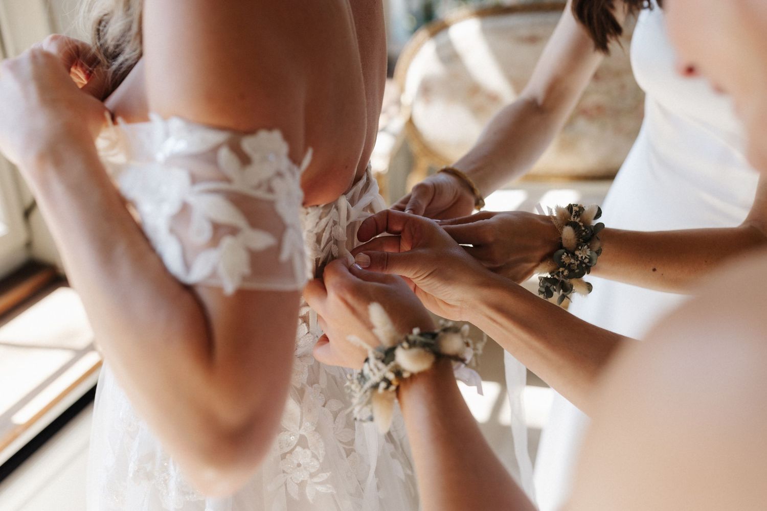 Bridesmaids helping fasten the back of a bride's lace wedding dress with floral wrist corsages.