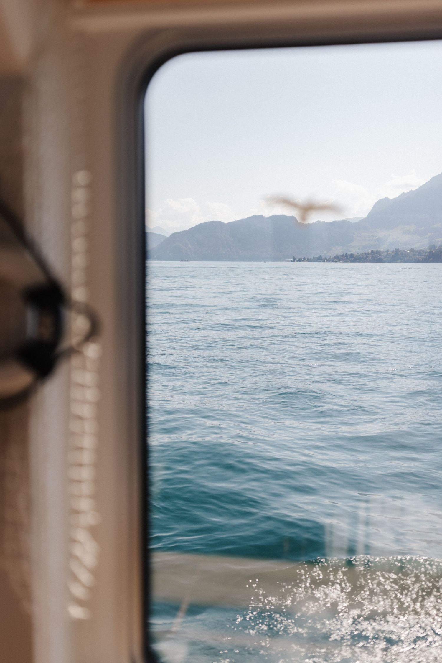 View through a boat window of blue lake water with mountains and a flying bird visible in the background.