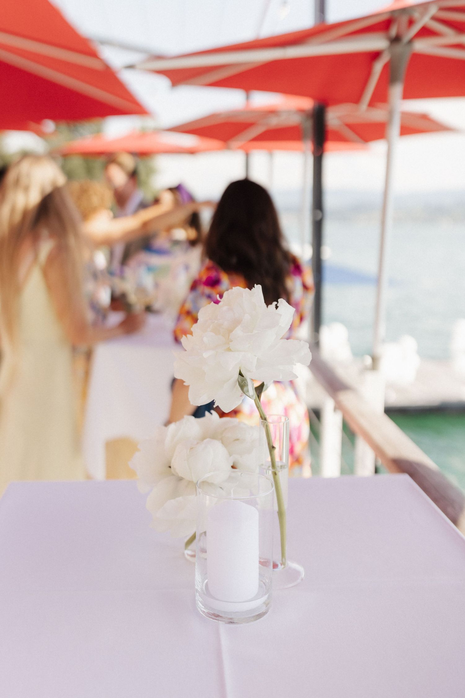 White peony flowers in a glass vase on a pink table at a waterfront restaurant with red umbrellas.