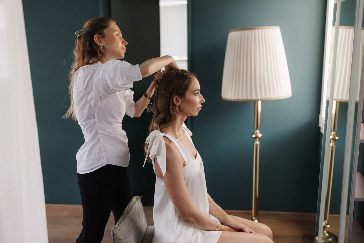Professional hairstylist working on seated bride's updo in elegant teal room.
