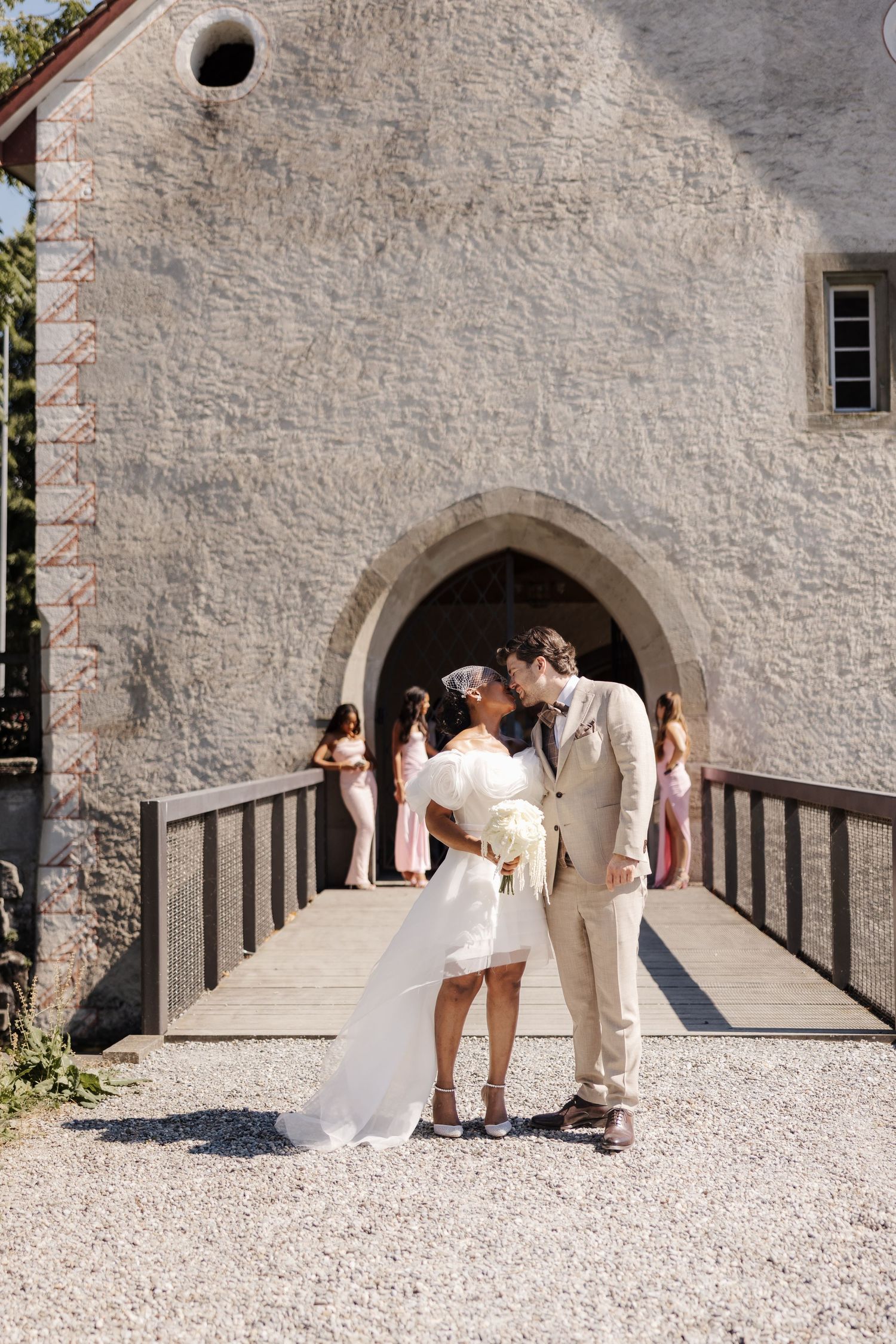 Newlywed interracial couple sharing a kiss on a bridge outside a historic stone church.