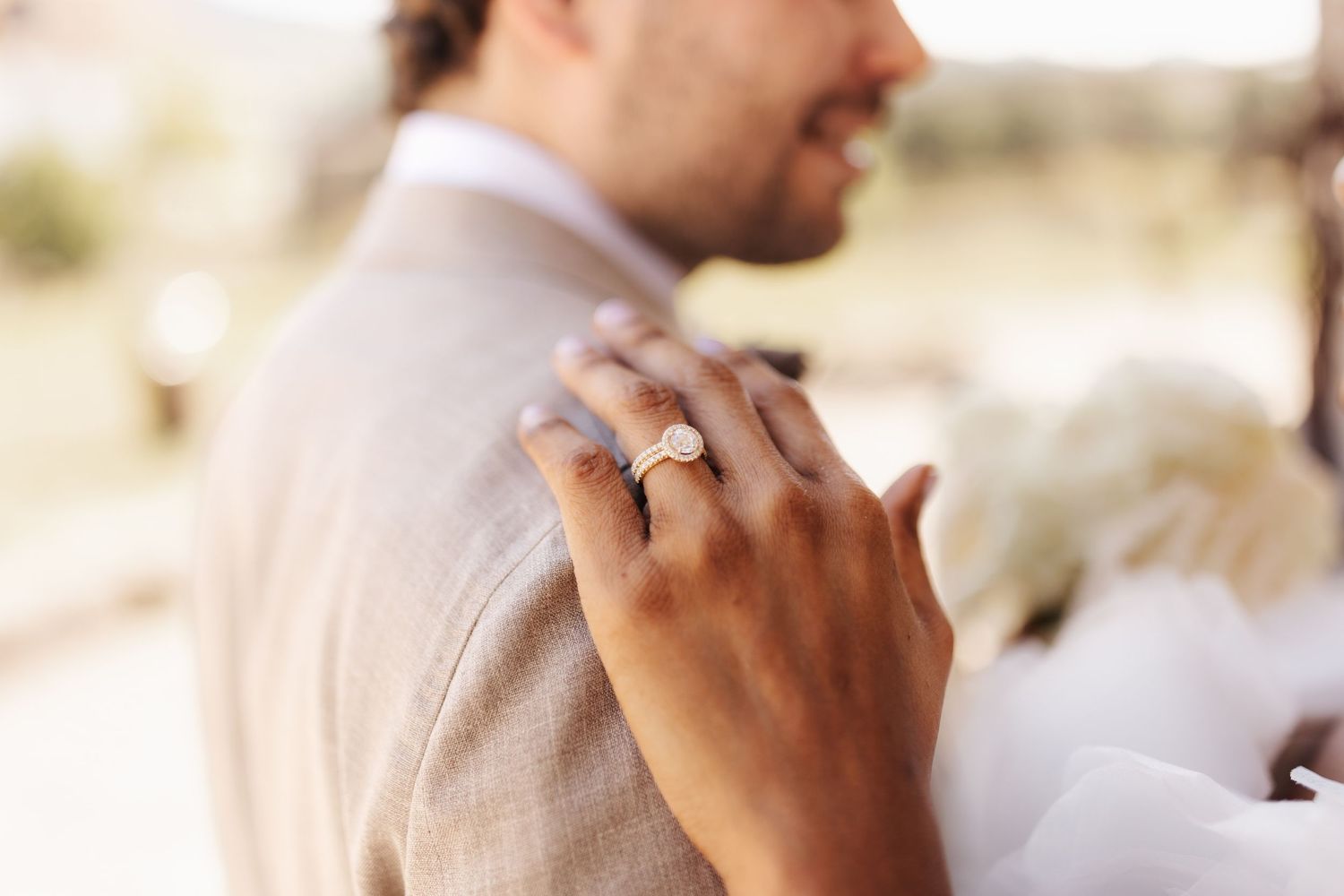 Close-up of bride's hand with diamond engagement ring resting on groom's beige suit jacket shoulder.