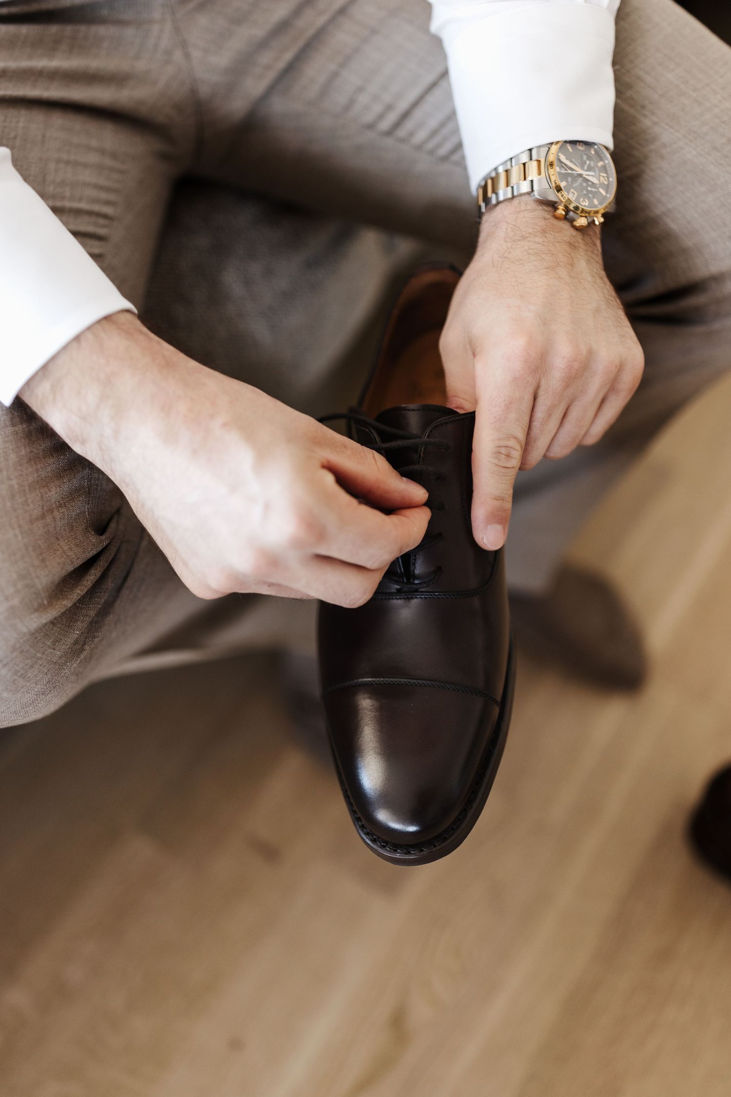 Groom in white shirt and gray trousers tying dark brown leather Oxford shoe laces.