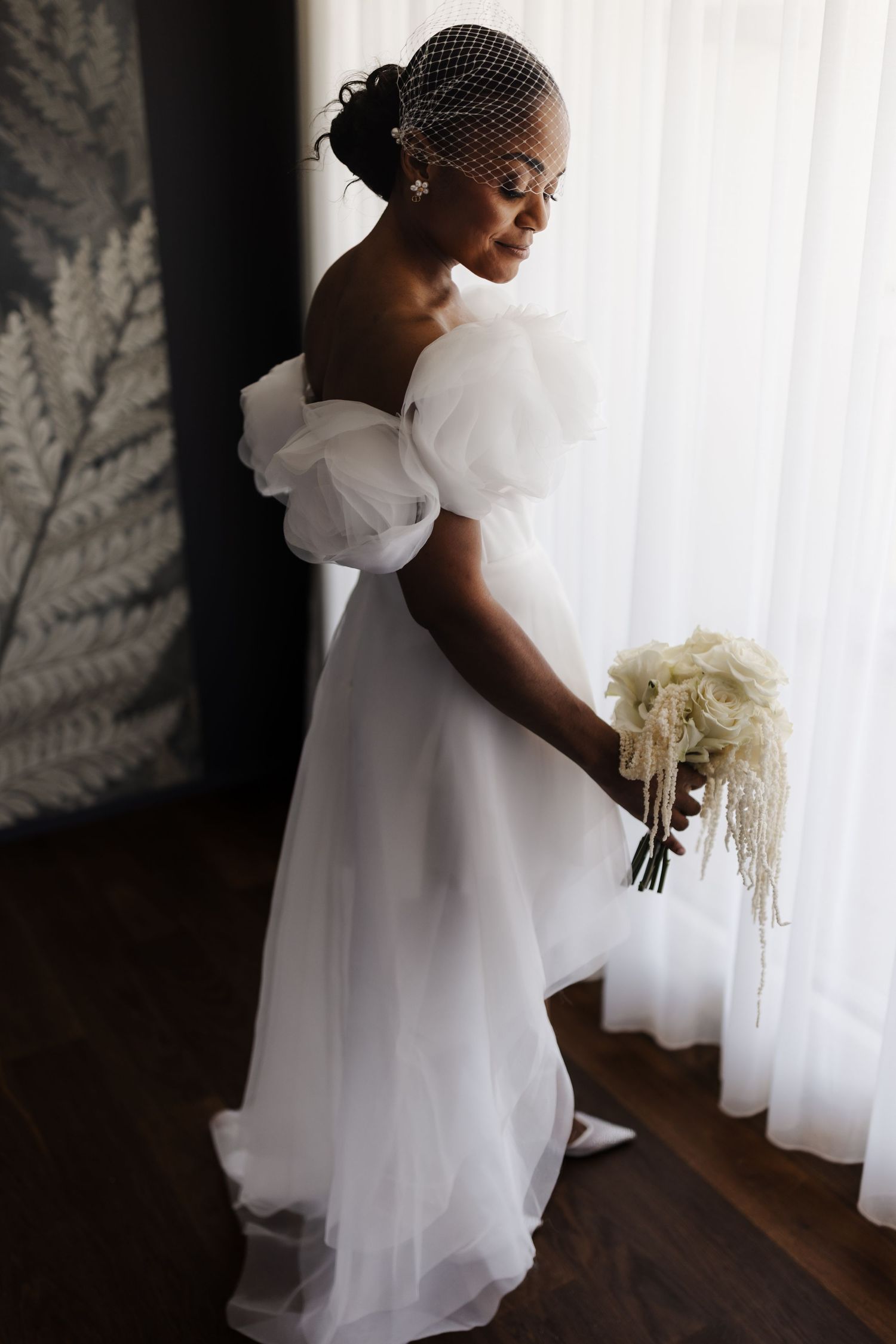 Black bride in white tulle off-shoulder gown and birdcage veil holds white bouquet by window.