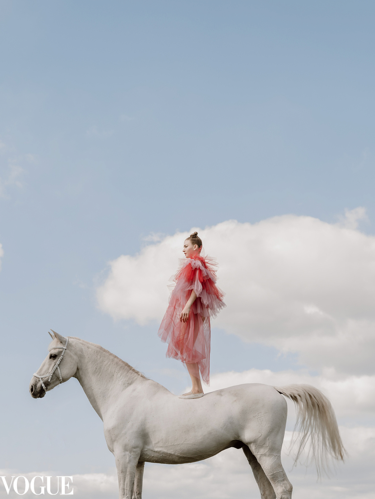 Model in red ruffled gown standing atop a white horse against a blue cloudy sky, Vogue editorial.