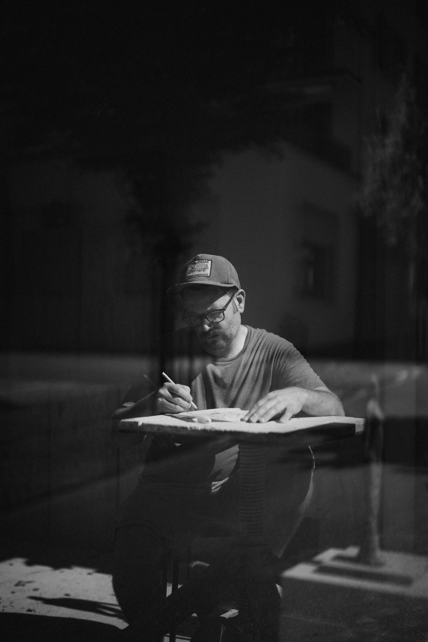 Black and white photo of bearded man in cap writing at desk outdoors at night.
