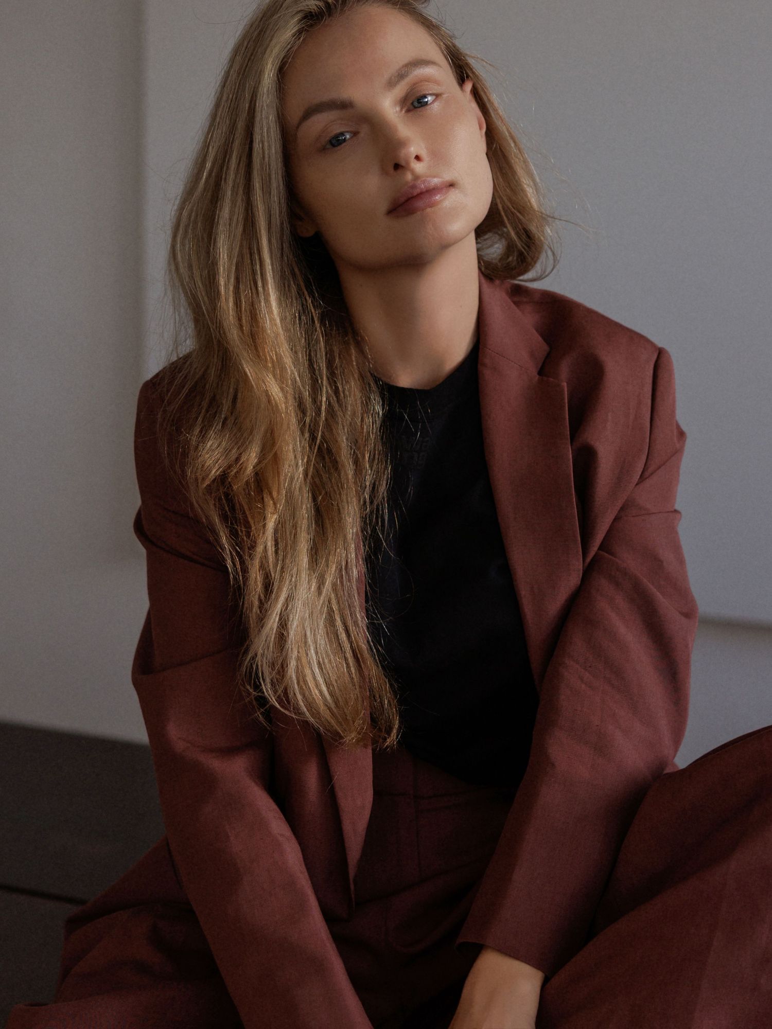 Woman with long blonde hair sits in a burgundy blazer and matching trousers over a black top, posing against a neutral wall.